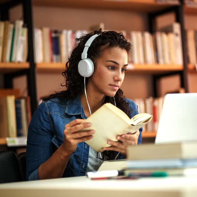 Woman wearing headphones, looking at laptop, and holding an open book