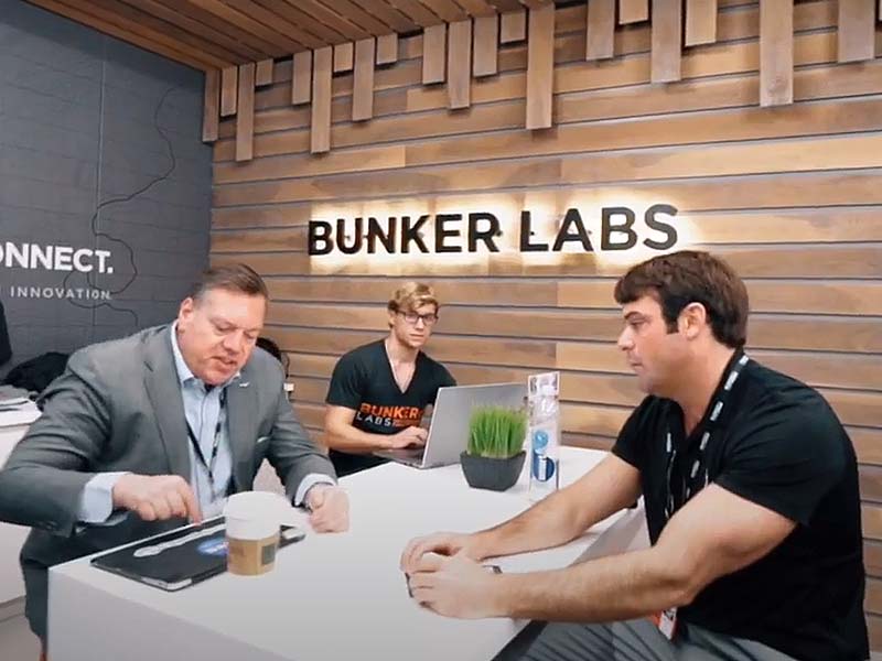 Three men at a table with the Bunker Labs sign on the wall behind them. The men are looking at each other while discussing something.