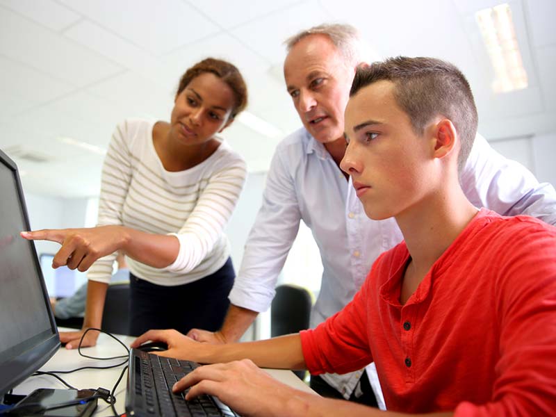 Two students, one boy, one girl, with their older male business teacher. The boy is sitting at a computer and the girl is pointing to the screen.