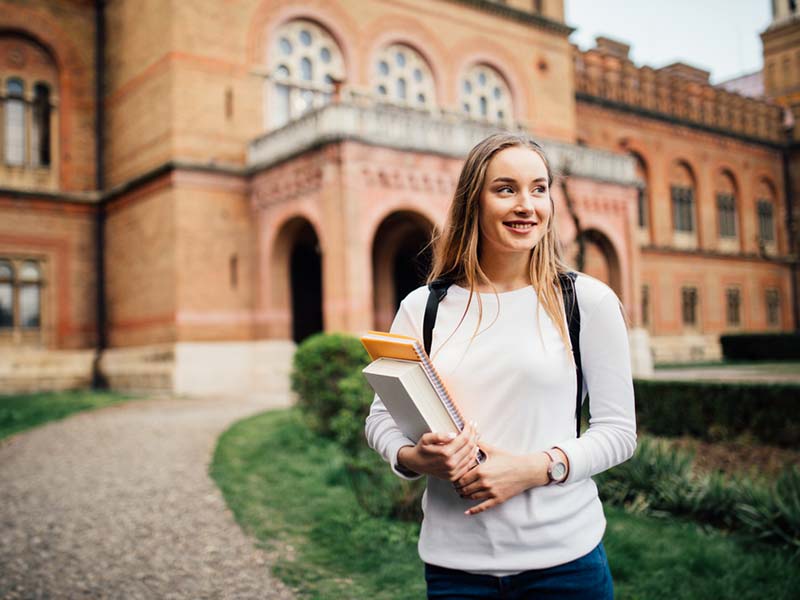 A young blonde woman holding some books and looking off into the distance. She is standing in front of a brick university building.