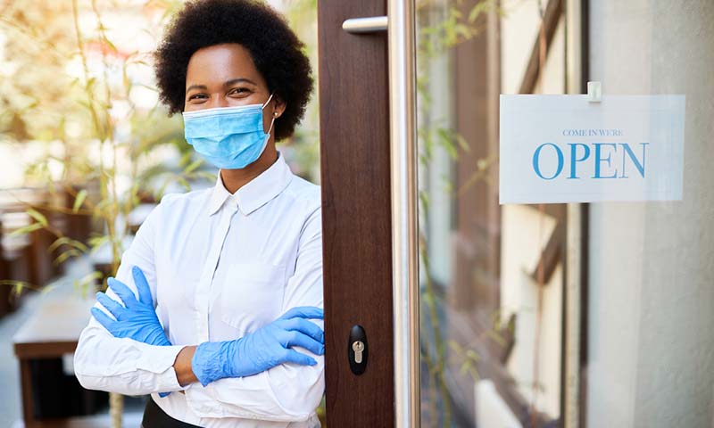 open-for-business A happy black woman with a mask and gloves is standing next to the door to her business with an open sign hanging in the window.