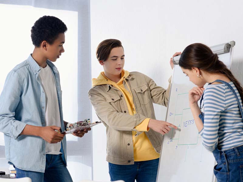 Three teens around a whiteboard. One of them is pointing at some plans that have been drawn on the board.