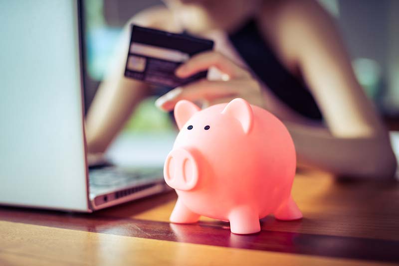 A piggy bank on a desk with a woman behind it on her laptop with her credit card ready to go.