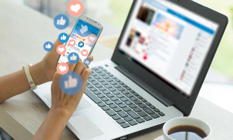 social-media A woman using her phone and laptop to engage with social media sites - heart and thumbs up icons are floating off her screen.
