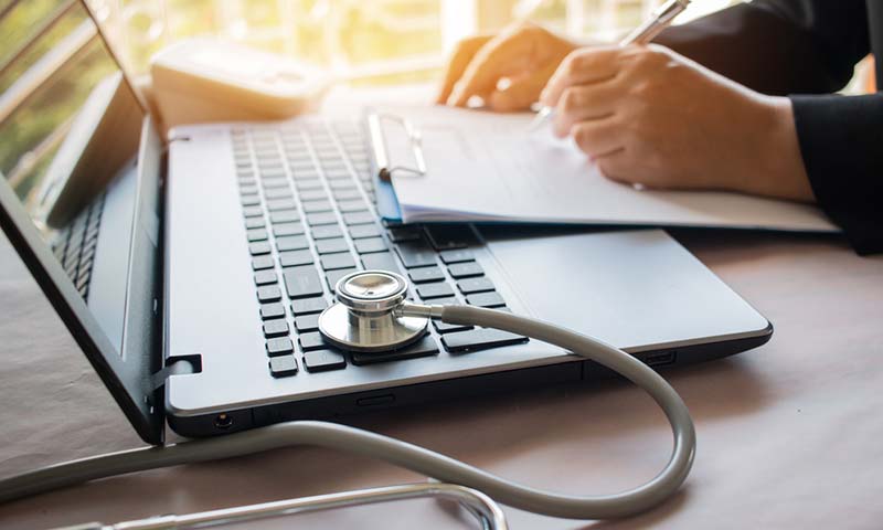payroll-checkup A laptop and someone doing paperwork. The laptop has a stethoscope on it.