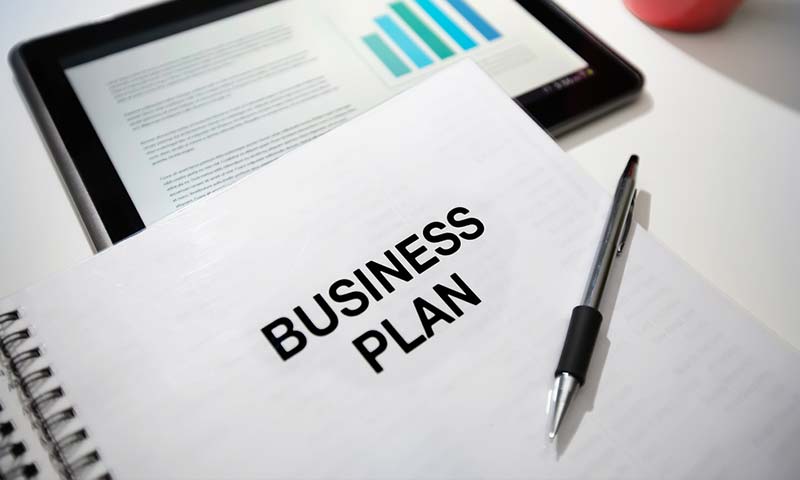 biz-plan A spiral notebook titled "Business Plan" sits on top of a digital tablet with a bar chart on it.