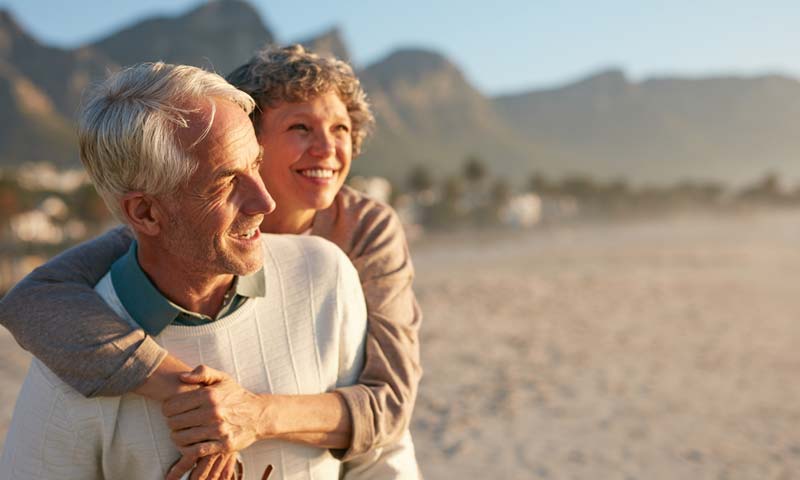 An elderly woman hugging her husband on a sandy beach