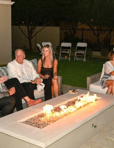 A group of mature people sitting around an outdoor gas firepit having cocktails.