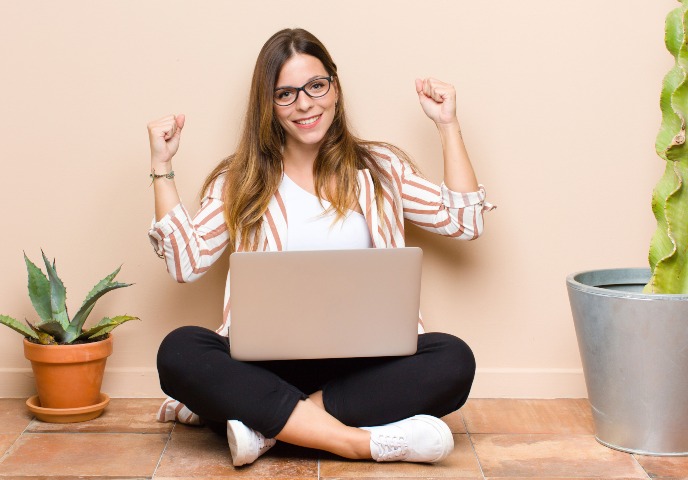 A female sitting cross-legged with laptop in her lap, hands raised as if she were cheering