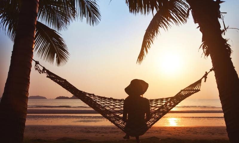 lady on a hammock between two palm trees on the beach looking out at the sunset