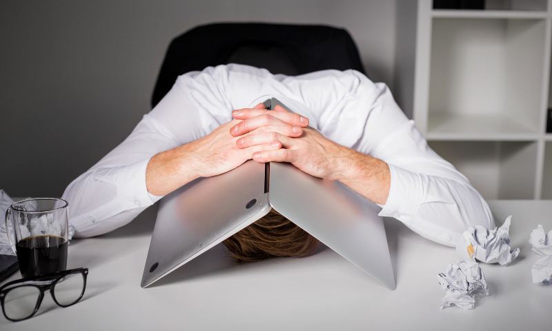 Business man sitting with head on desk, open laptop covering his head
