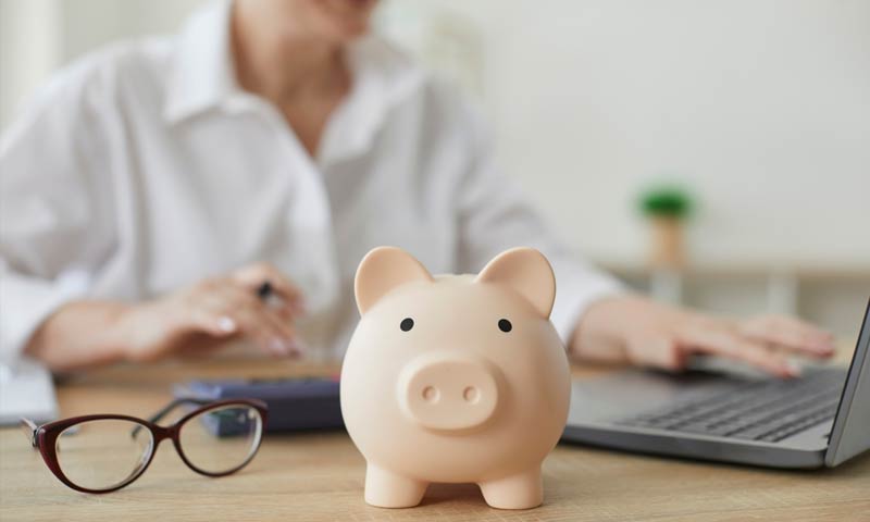 Piggy bank on a desk with a woman behind it using a laptop and calculator.