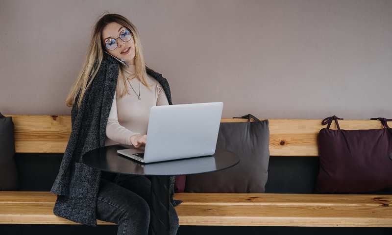 Young lady in glasses sitting on wooden bench working on laptop and talking on her phone.
