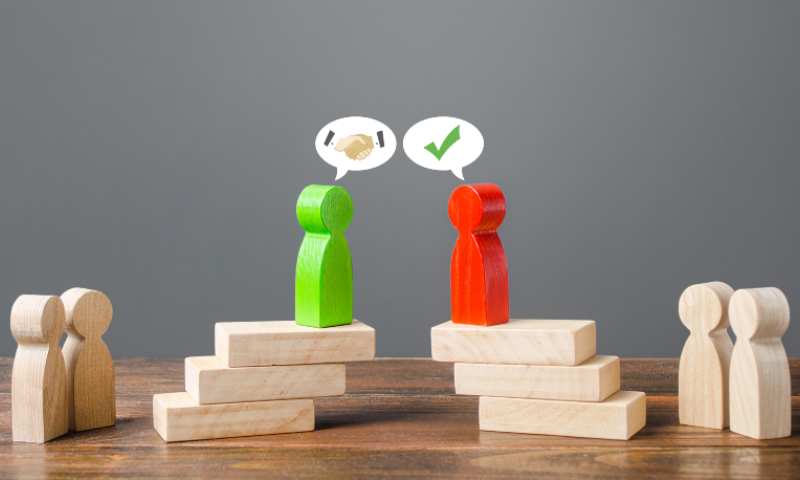 Wooden blocks stacked with wooden figures, green and red figure facing one another, speech bubbles above