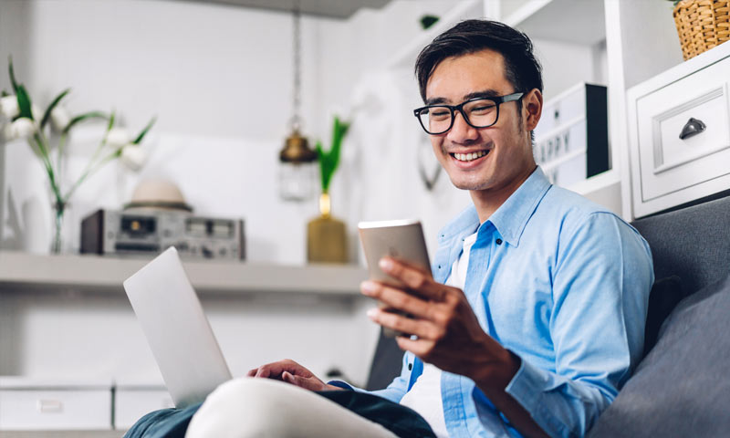 Smiling Asian man wearing a blue business shirt looking at laptop and smart phone